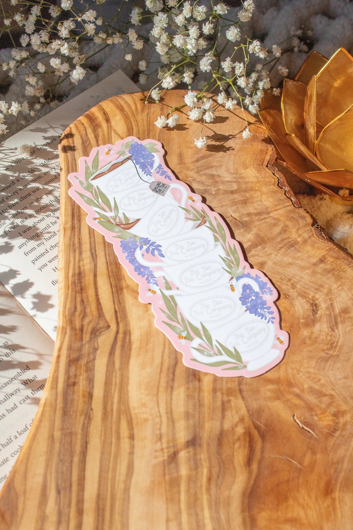 Bridgeton Inspired Die-Cut Bookmark for Regency Era Romance Readers. 

A regency era romance tea cup stack design with ivory tea cups that say "The Ton Tea Co. London 1819" surrounded by wisteria on a light pink background. The bookmark is sitting on a wood cutting board, on an open book surrounded by little white flowers. 