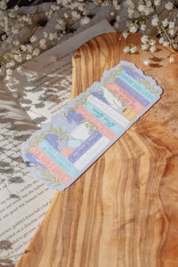 Bridgeton Inspired Die-Cut Bookmark for Regency Era Romance Readers. 

A regency era romance bookshelf design in light pinks, blues, violet, and ivory surrounded by wisteria on a lilac background. The bookmark is sitting on a wood cutting board, on an open book surrounded by little white flowers. 