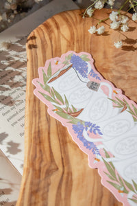 Bridgeton Inspired Die-Cut Bookmark for Regency Era Romance Readers. 

A regency era romance tea cup stack design with ivory tea cups that say "The Ton Tea Co. London 1819" surrounded by wisteria on a light pink background. The bookmark is sitting on a wood cutting board, on an open book surrounded by little white flowers. 
