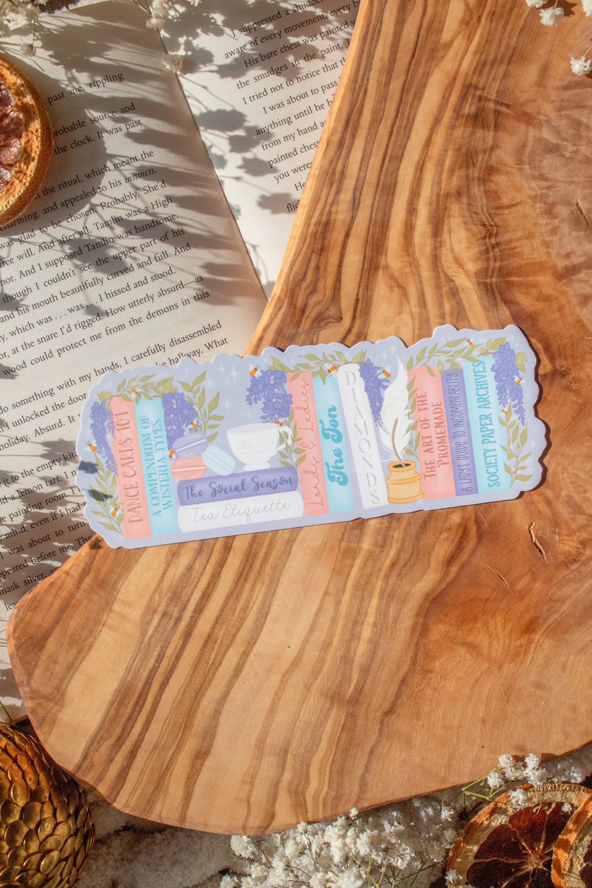 Bridgeton Inspired Die-Cut Bookmark for Regency Era Romance Readers. 

A regency era romance bookshelf design in light pinks, blues, violet, and ivory surrounded by wisteria on a lilac background. The bookmark is sitting on a wood cutting board, on an open book surrounded by little white flowers. 