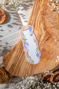 Bridgeton Inspired Die-Cut Bookmark for Regency Era Romance Readers. 

A regency era romance tea cup stack design with ivory tea cups that say "The Ton Tea Co. London 1819" surrounded by wisteria on a light pink background. The bookmark is sitting on a wood cutting board, on an open book surrounded by little white flowers. 