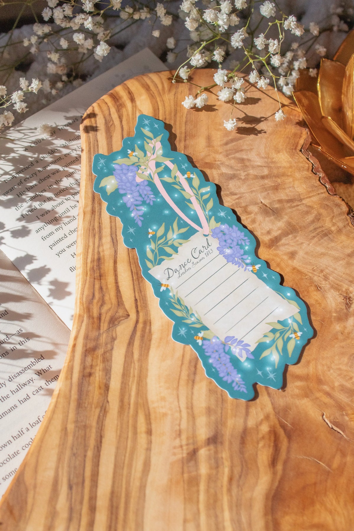 Bridgeton Inspired Die-Cut Bookmark for Regency Era Romance Readers. 

This bookmark is a regency style dance card on a teal-blue background surrounded by wisteria, little bees, and stars. The Bookmark is on a olive wood cutting board, on an open book and surrounded by little white flowers.