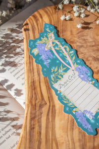Bridgeton Inspired Die-Cut Bookmark for Regency Era Romance Readers. 

This bookmark is a regency style dance card on a teal-blue background surrounded by wisteria, little bees, and stars. The Bookmark is on a olive wood cutting board, on an open book and surrounded by little white flowers.