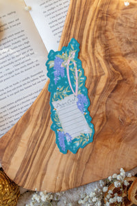 Bridgeton Inspired Die-Cut Bookmark for Regency Era Romance Readers. 

This bookmark is a regency style dance card on a teal-blue background surrounded by wisteria, little bees, and stars. The Bookmark is on a olive wood cutting board, on an open book and surrounded by little white flowers. 