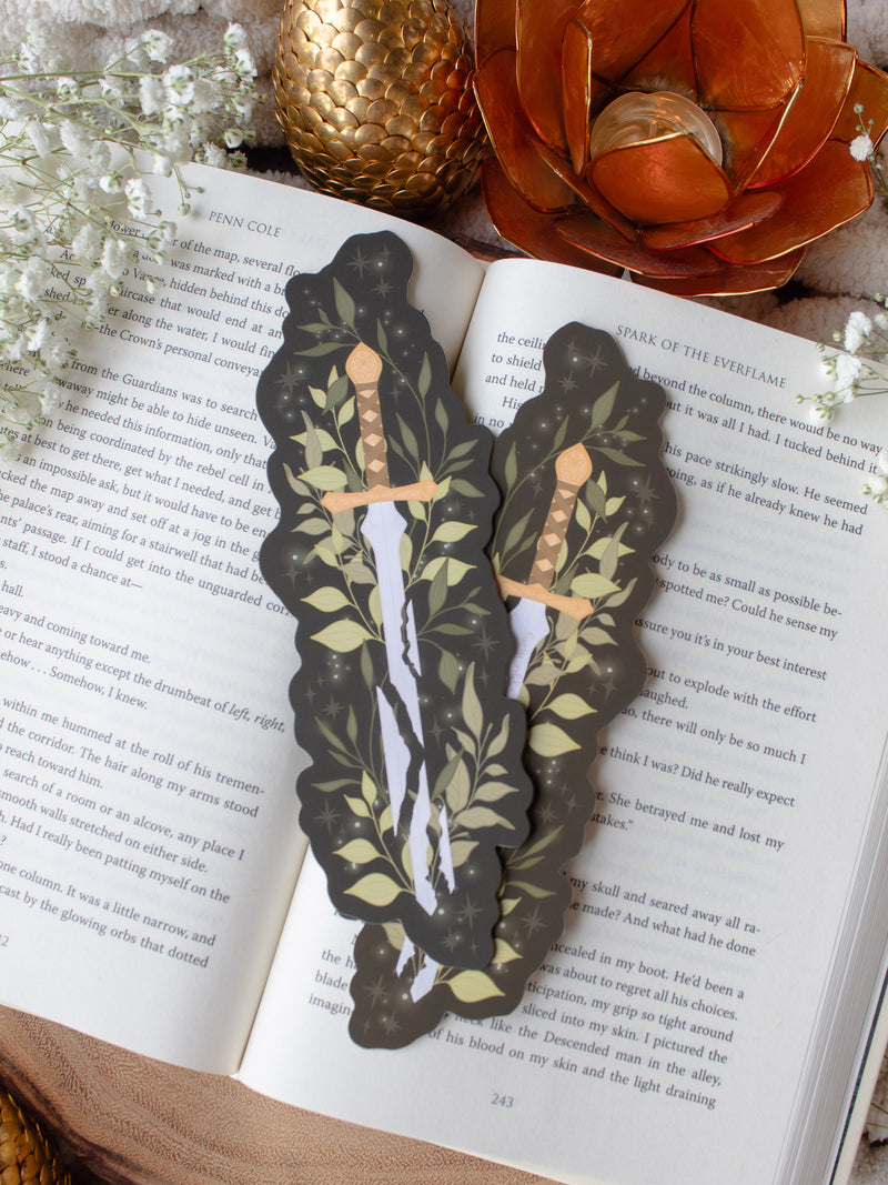Broken Fantasy Sword Die-Cut Bookmark on an open book with a gold metal dragon egg, baby's breath, and an orange lotus tea light holder in the background. 