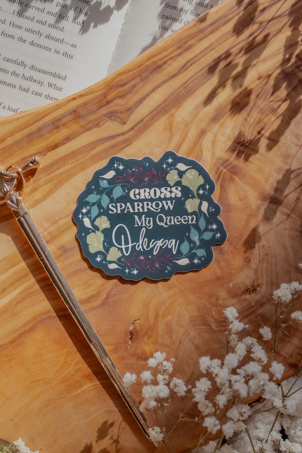 Shield of Sparrow bookish waterproof vinyl sticker inspired by Devney Perry series. This sticker circle-ish in shape, with a dark teal background and the words "Cross, Sparrow, My Queen, Odessa" on it. Each on a different line with foliage surrounding them. 

The sticker is sitting on an olive wood cutting board, on top of an open book, with little white flowers 
