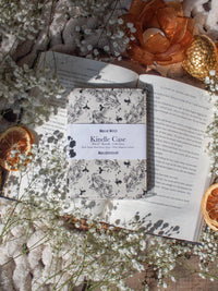 Ivory Swords Fantasy Reader Dragon Kindle Case. It has an ivory background with deep charcoal dragons, swords, crowns, and foliage all over it. This is a full cover kindle case with a leatherette outside, a kick-stand, card-slot, and hand-strap. 