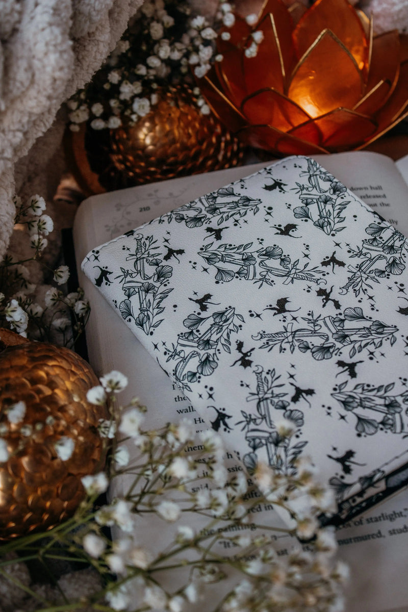 Ivory Swords Fantasy Reader Dragon Kindle Case. It has an ivory background with deep charcoal dragons, swords, crowns, and foliage all over it. This is a full cover kindle case with a leatherette outside, a kick-stand, card-slot, and hand-strap. 