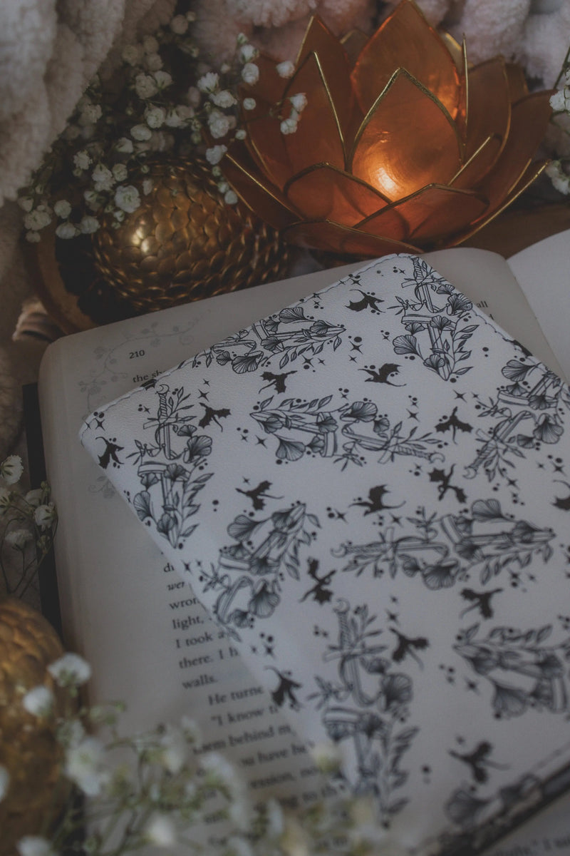 Ivory Swords Fantasy Reader Dragon Kindle Case. It has an ivory background with deep charcoal dragons, swords, crowns, and foliage all over it. This is a full cover kindle case with a leatherette outside, a kick-stand, card-slot, and hand-strap. 