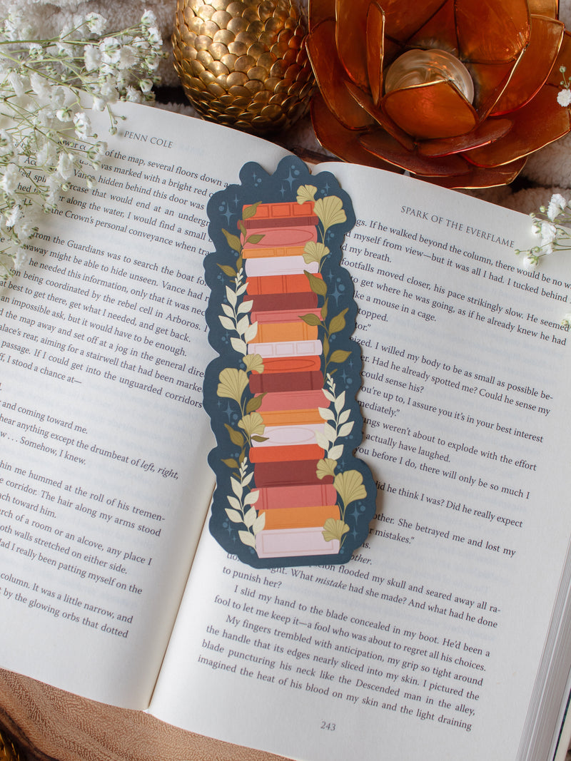 This moody toned fall autumn bookstack bookmark is a bookstack in warm tones, like mustard yellow, cream, coral, and burnt orange on a deep teal background. 