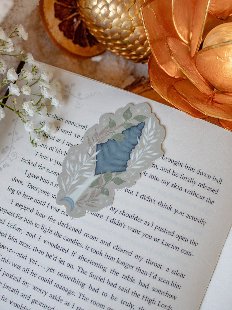 This bookish When The Moon Hatched dragonscale dagger sticker is inspired by the Moonfall series by Sarah A Parker. The image shows the sticker on an open book, with the book placed on a fuzzy neutral colored blanket, a faux golden dragon egg, orange slices, and an orange lotus shaped tea light holder all surrounded by little white flowers