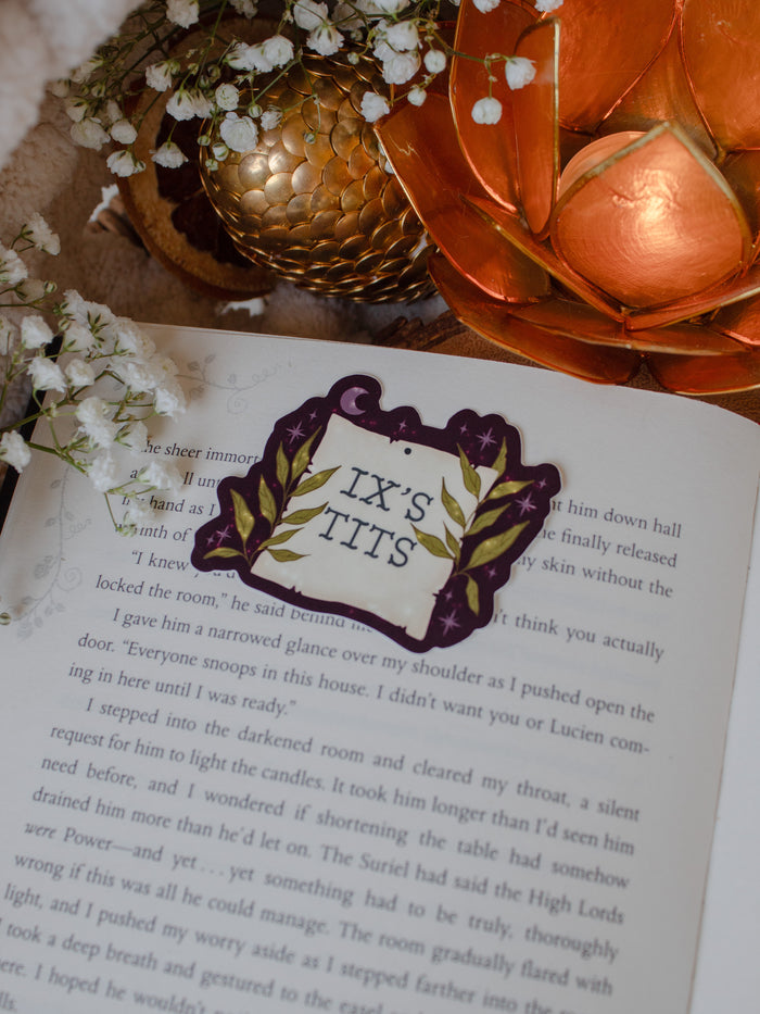 This officially licensed Serpent and The Wings of Night sticker from the Crowns of Nyaxia Series by Carissa Broadbent features the text "Ix's Tits" on a note that looks like weathered parchment on a deep maroon background with foliage, little stars and magic bubbles surrounding it. 