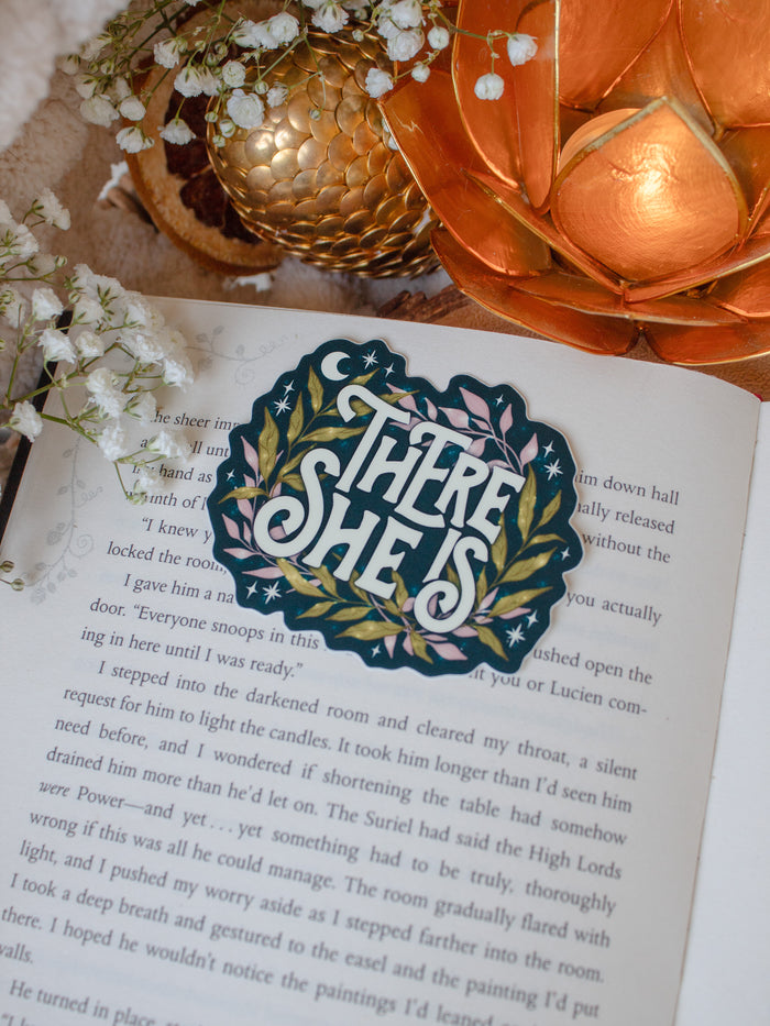 This officially licensed Serpent and The Wings of Night Sticker features the text "There She Is" in a cream text on a deep teal background with pink and green foliage surrounding it with little stars and magic bubbles. 