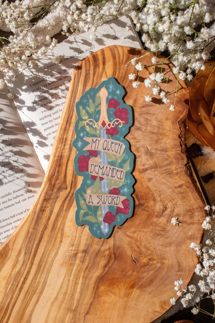 Bookmark inspired by the novel by Devney Perry. This bookmark is teal with a sword surrounded by foliage and the text "My Queen Demanded a Sword" on a banner wrapped around the blade.

The bookmark is laid on an olive wood cutting board, sitting on an open book with little white flowers surrounding it.