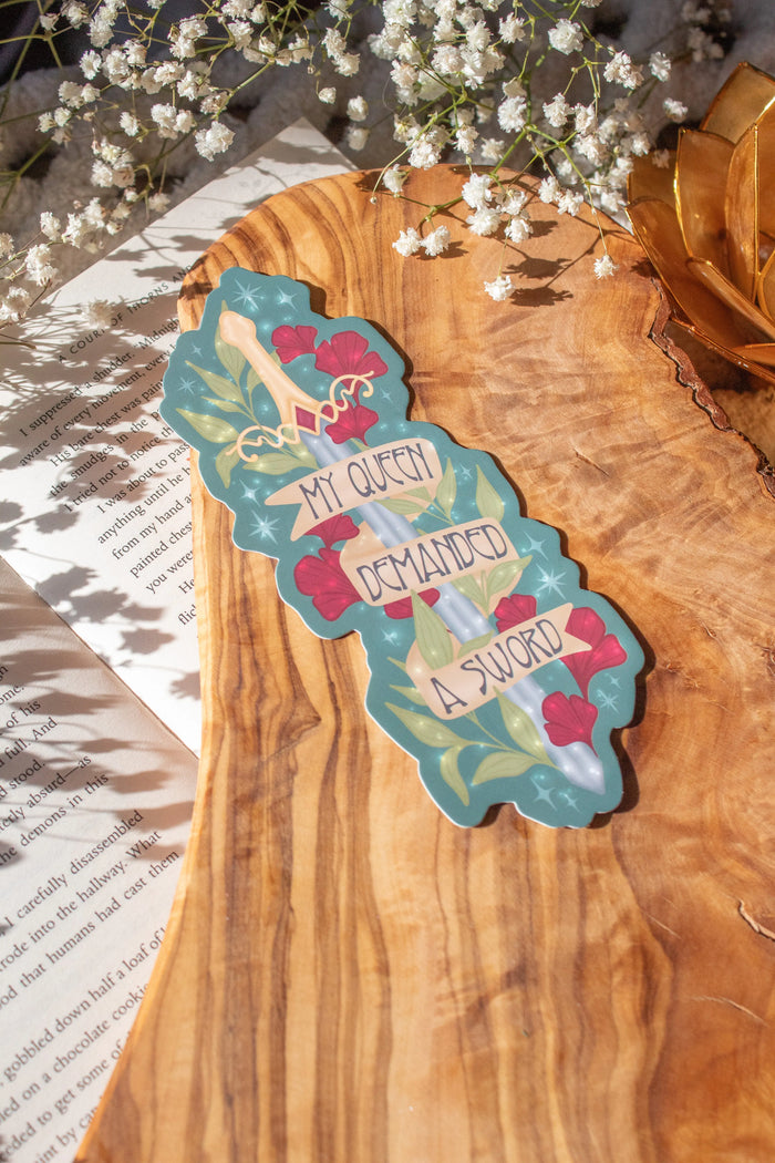Bookmark inspired by the novel by Devney Perry. This bookmark is teal with a sword surrounded by foliage and the text "My Queen Demanded a Sword" on a banner wrapped around the blade.

The bookmark is laid on an olive wood cutting board, sitting on an open book with little white flowers surrounding it.