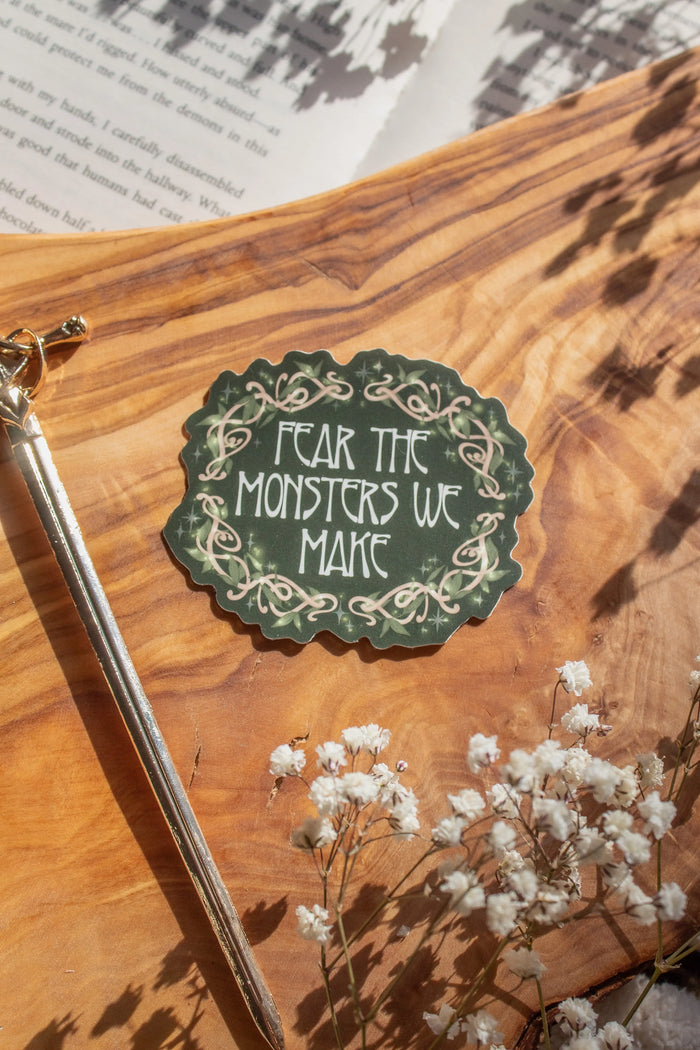 Shield of Sparrow bookish waterproof vinyl sticker inspired by Devney Perry series. This sticker is dark green, oval-ish in shape with "Fear the Monsters We Make" on it in an art nouveau font, and wood vines drawn around it in a Nordic Viking-style where they are woven together, foliage is intertwined with the wood vines and little magical looking bubbles surround them both. 

The sticker is sitting on an olive wood cutting board, on top of an open book, with little white flowers surrounding it.