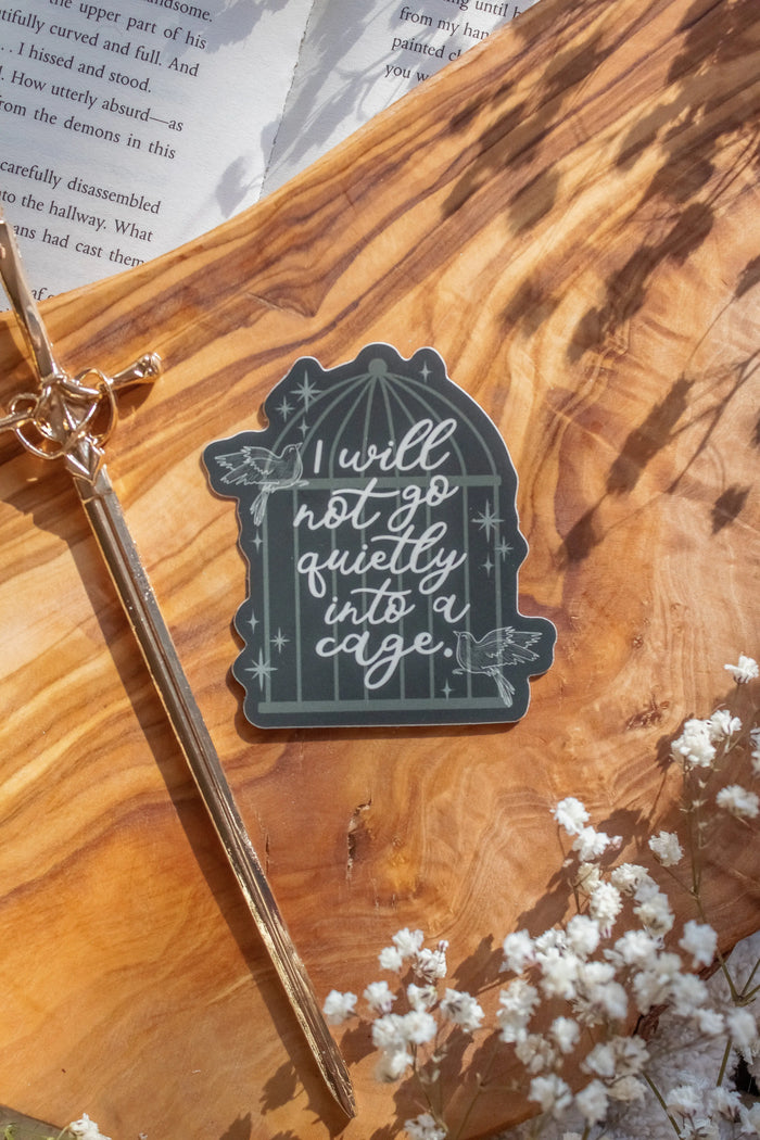 Shield of Sparrow bookish waterproof vinyl sticker inspired by Devney Perry series. This sticker is dark green, with a cage image on it with the words "I will not go quietly into a cage" on it in a pretty white script and little sparrows flying around it. 

The sticker is sitting on an olive wood cutting board, on top of an open book, with little white flowers surrounding it.