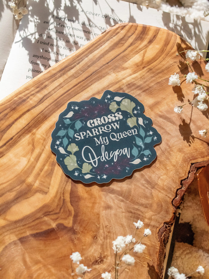 Shield of Sparrow bookish waterproof vinyl sticker inspired by Devney Perry series. This sticker circle-ish in shape, with a dark teal background and the words "Cross, Sparrow, My Queen, Odessa" on it. Each on a different line with foliage surrounding them. 

The sticker is sitting on an olive wood cutting board, on top of an open book, with little white flowers 
