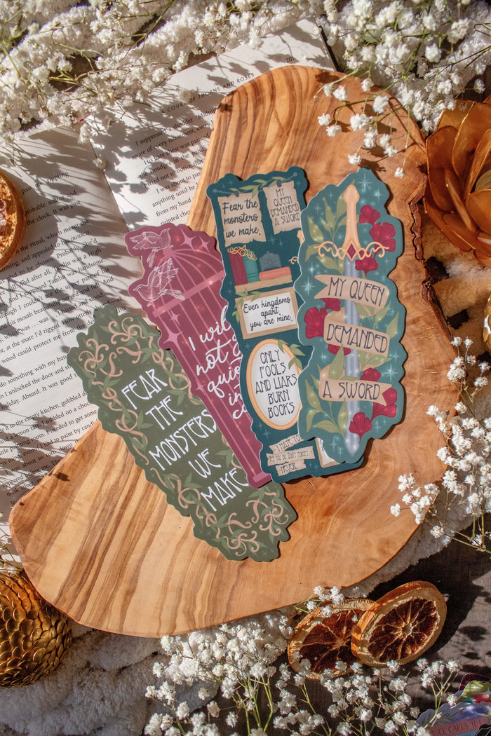 4 Shield of Sparrows Die-Cut Bookmarks, inspired by the book by Devney Perry. The bookmarks are laid on an olive wood cutting board, sitting on an open book with little white flowers, orange slices, and a gold dragon egg surrounding them.