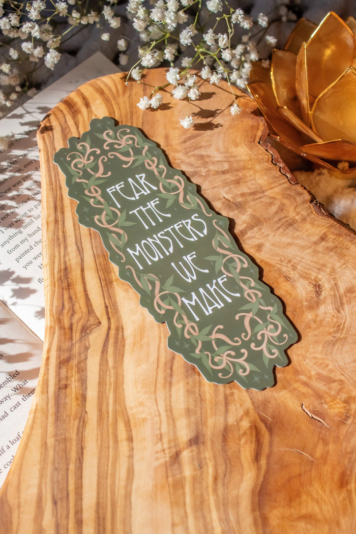 Bookmark inspired by the novel by Devney Perry. This bookmark is dark green with wood looking vines intertwined with foliage surrounding the words "Fear The Monsters We Make". 
The bookmark is laid on an olive wood cutting board, sitting on an open book with little white flowers surrounding it.