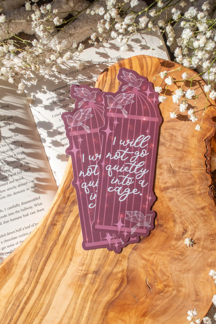 A Shield of Sparrows Die-Cut Bookmark inspired by the novel by Devney Perry. This bookmark is maroon with a cage on it, and little sparrows flying around it along with little stars and magic looking bubbles. It also has the text "I will not go quietly into a cage" on it in a pretty white script.

The bookmark is laid on an olive wood cutting board, sitting on an open book with little white flowers surrounding it.