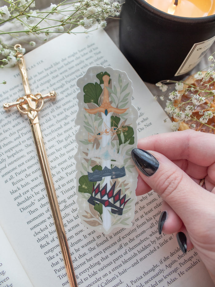 Spark of The Everflame Diem's Crown Sword Sticker, Lunmos Crown, Montios Crown, Fortos Crown, Umbros Crown. From The Kindred's Curse Saga by Penn Cole. This officially licensed sticker is a sword on a light tan background, through the forementioned crowns. Surrounded by foliage. 