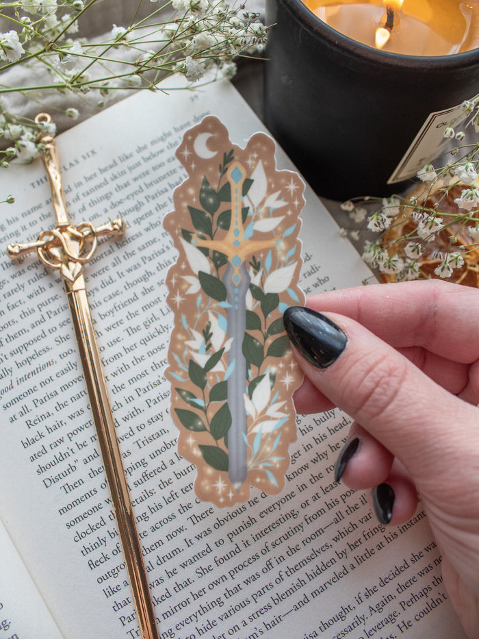 Spark of the Everflame Sword of Corbois waterproof and dishwasher safe vinyl sticker. From the Kindred's Curse Saga by Penn Cole. This officially licensed sticker is a dark metal sword, surrounded by foliage, on a burn orange background. 
