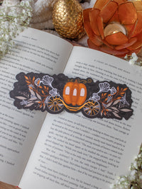 This spooky / halloween fairytale inspired midnight pumpkin carriage ride bookmark features a pumpkin shaped carriage with wheels off both sides. Surrounded by orange, tan, and white foliage that are covered in spider webs. 