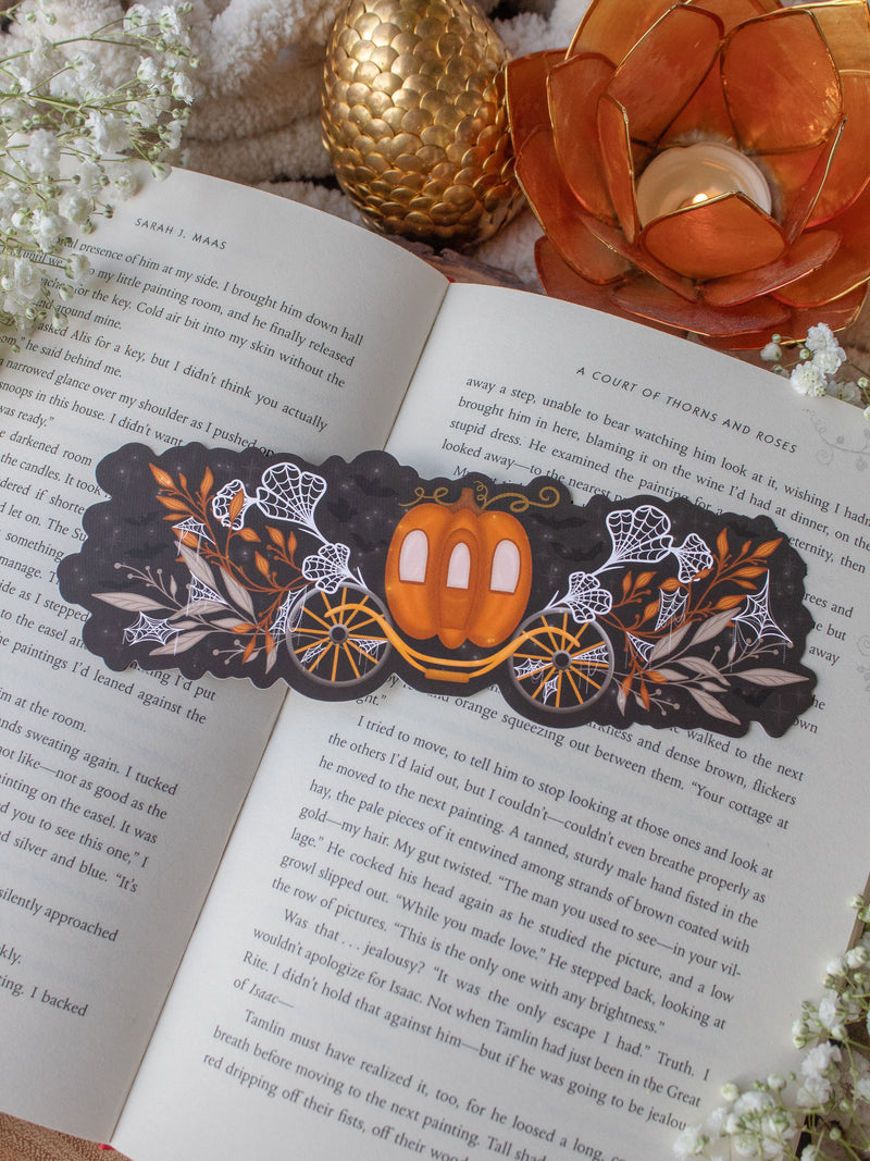 This spooky / halloween fairytale inspired midnight pumpkin carriage ride bookmark features a pumpkin shaped carriage with wheels off both sides. Surrounded by orange, tan, and white foliage that are covered in spider webs. 
