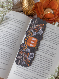 This spooky / halloween fairytale inspired midnight pumpkin carriage ride bookmark features a pumpkin shaped carriage with wheels off both sides. Surrounded by orange, tan, and white foliage that are covered in spider webs. 