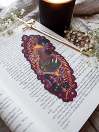 This fantasy spooky / halloween sleeping dragon elixir bookmark features a little black dragon curled up in a potion bottle with their tale sticking out the top. Surrounded by orange, black, and light purple foliage  covered in spider webs, with little stars and magic bubbles floating around. On a maroon/ deep purple background. 