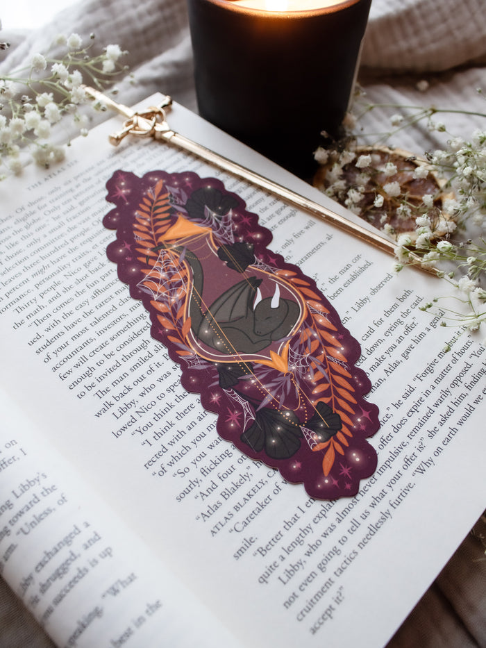 This fantasy spooky / halloween sleeping dragon elixir bookmark features a little black dragon curled up in a potion bottle with their tale sticking out the top. Surrounded by orange, black, and light purple foliage  covered in spider webs, with little stars and magic bubbles floating around. On a maroon/ deep purple background. 