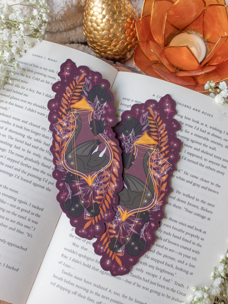 This fantasy spooky / halloween sleeping dragon elixir bookmark features a little black dragon curled up in a potion bottle with their tale sticking out the top. Surrounded by orange, black, and light purple foliage  covered in spider webs, with little stars and magic bubbles floating around. On a maroon/ deep purple background. 