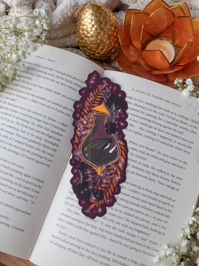 This fantasy spooky / halloween sleeping dragon elixir bookmark features a little black dragon curled up in a potion bottle with their tale sticking out the top. Surrounded by orange, black, and light purple foliage  covered in spider webs, with little stars and magic bubbles floating around. On a maroon/ deep purple background. 