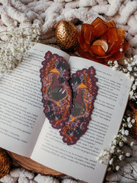 This fantasy spooky / halloween sleeping dragon elixir bookmark features a little black dragon curled up in a potion bottle with their tale sticking out the top. Surrounded by orange, black, and light purple foliage  covered in spider webs, with little stars and magic bubbles floating around. On a maroon/ deep purple background. 
