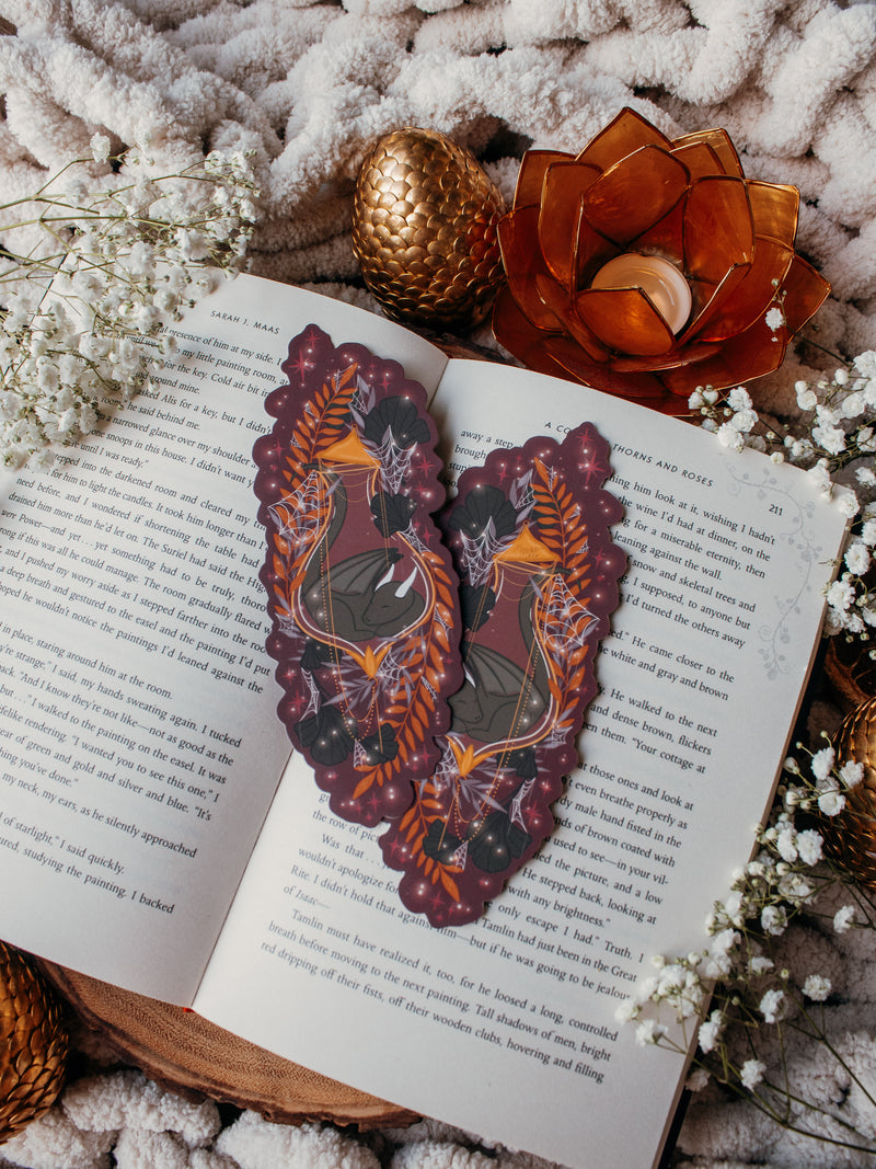 This fantasy spooky / halloween sleeping dragon elixir bookmark features a little black dragon curled up in a potion bottle with their tale sticking out the top. Surrounded by orange, black, and light purple foliage  covered in spider webs, with little stars and magic bubbles floating around. On a maroon/ deep purple background. 
