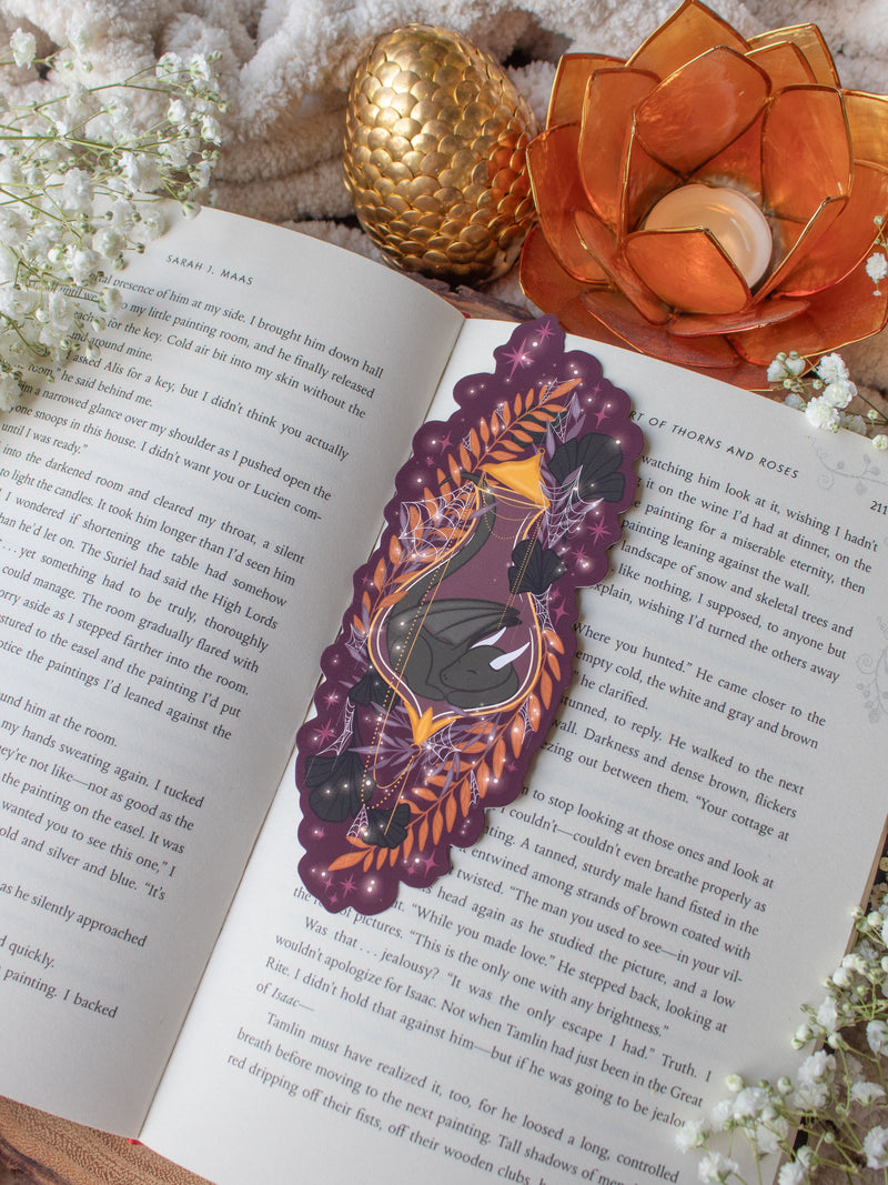This fantasy spooky / halloween sleeping dragon elixir bookmark features a little black dragon curled up in a potion bottle with their tale sticking out the top. Surrounded by orange, black, and light purple foliage  covered in spider webs, with little stars and magic bubbles floating around. On a maroon/ deep purple background. 