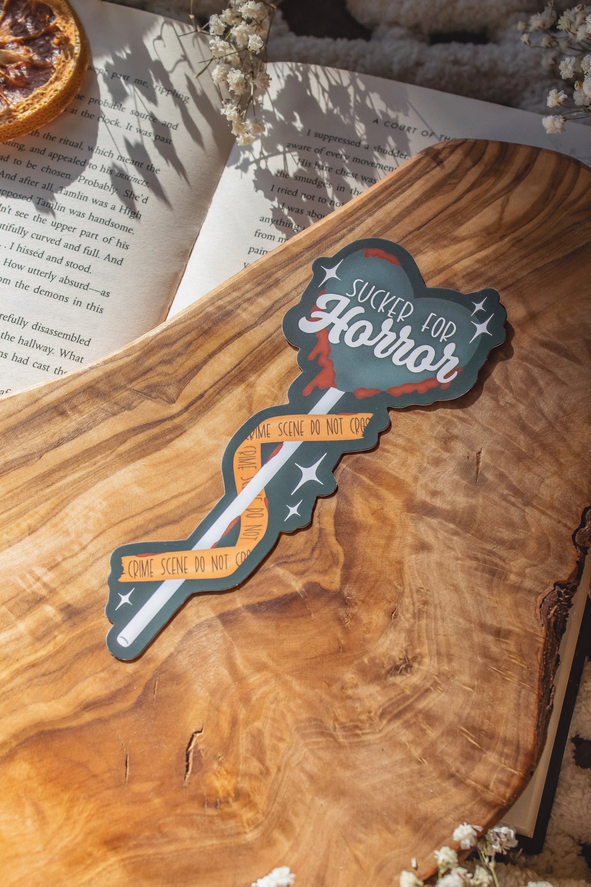 This Sucker for Horror Die-Cut Bookmark is for all my mystery loving book babes who love to delve into a good thriller. 

It is a dark grey heart shaped sucker with blood dripping off of it, wrapped in crime scene tape and surrounded by stars on a darker grey background.


The bookmark is sitting on an olive wood cutting board, on an open book, with little white flowers surrounding it.
