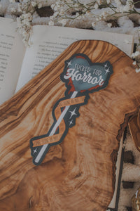 This Sucker for Horror Die-Cut Bookmark is for all my mystery loving book babes who love to delve into a good thriller. 

It is a dark grey heart shaped sucker with blood dripping off of it, wrapped in crime scene tape and surrounded by stars on a darker grey background.


The bookmark is sitting on an olive wood cutting board, on an open book, with little white flowers surrounding it.