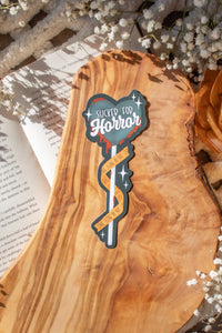 This Sucker for Horror Die-Cut Bookmark is for all my mystery loving book babes who love to delve into a good thriller. 

It is a dark grey heart shaped sucker with blood dripping off of it, wrapped in crime scene tape and surrounded by stars on a darker grey background.


The bookmark is sitting on an olive wood cutting board, on an open book, with little white flowers surrounding it.