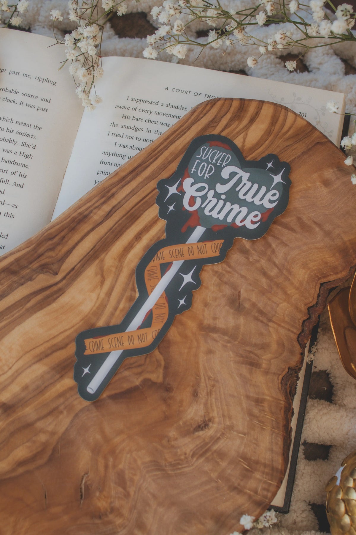 This Sucker for True Crime Die-Cut Bookmark is for all my mystery loving book babes who love to delve into a good cold case file. 

It is a dark grey heart shaped sucker with blood dripping off of it, wrapped in crime scene tape and surrounded by stars on a darker grey background.


The bookmark is sitting on an olive wood cutting board, on an open book, with little white flowers surrounding it.
