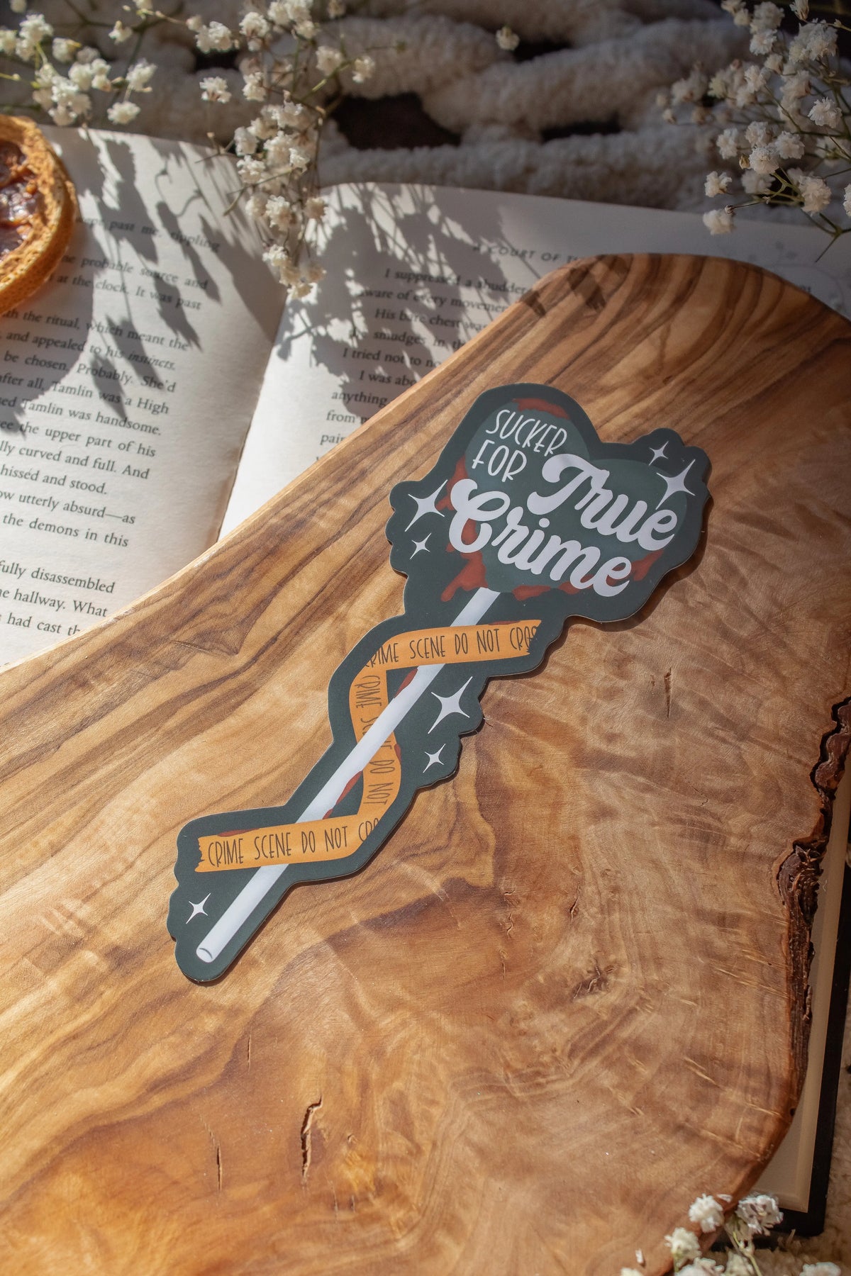 This Sucker for True Crime Die-Cut Bookmark is for all my mystery loving book babes who love to delve into a good cold case file. 

It is a dark grey heart shaped sucker with blood dripping off of it, wrapped in crime scene tape and surrounded by stars on a darker grey background.


The bookmark is sitting on an olive wood cutting board, on an open book, with little white flowers surrounding it.
