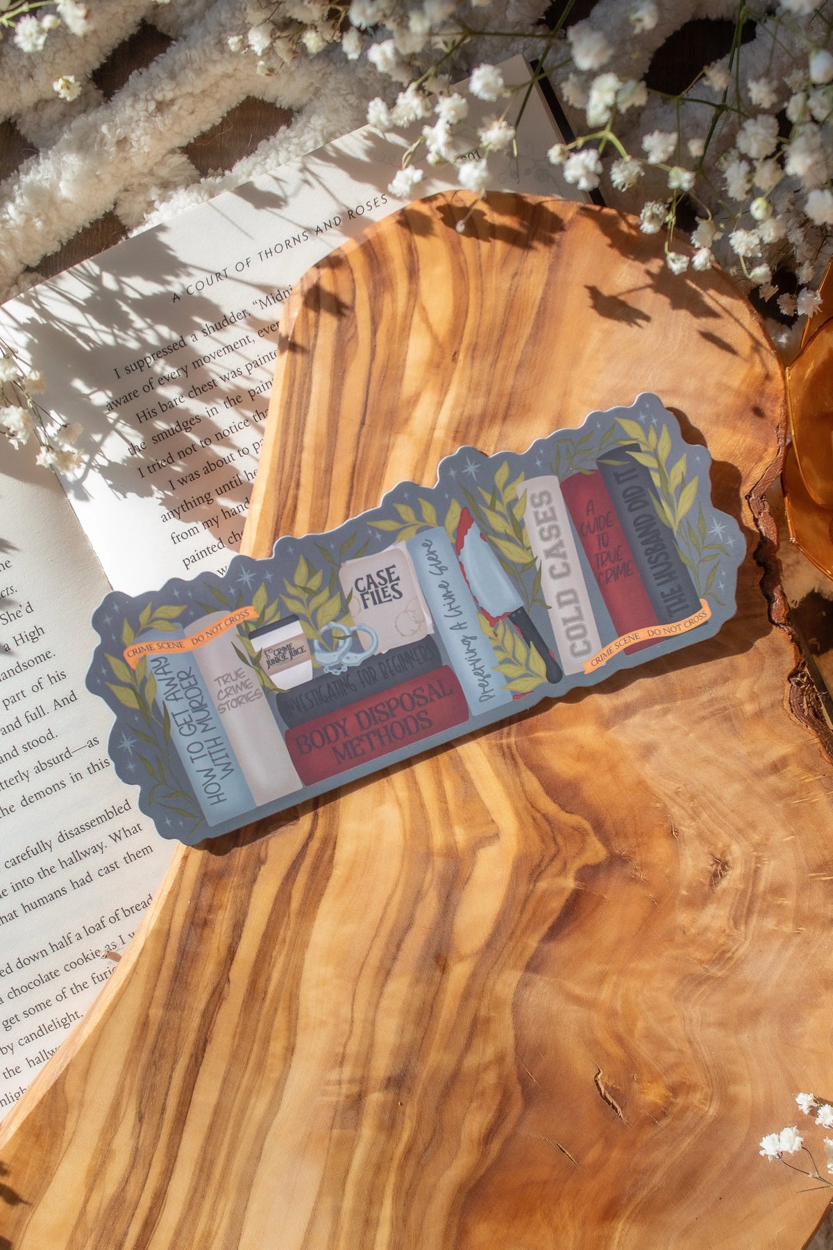 This True Crime Reader Bookshelf Die-Cut Bookmark is for all my mystery loving book babes who love to delve into a good cold case file. 

This bookmark shows a bookshelf with red, black, grey, and taupe books surrounded by crime scene tape, hand cuffs, coffee, case files, and a bloody knife. 

The bookmark is sitting on an olive wood cutting board, on an open book, with little white flowers surrounding it. 