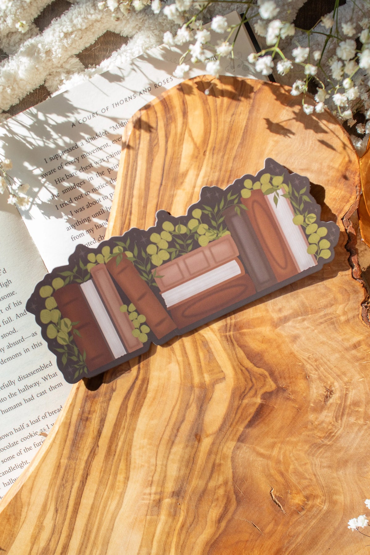 This Vintage  Bookshelf Die-Cut Bookmark is for all my classical book loving babes who love to delve into a classic.

This bookmark shows a bookshelf with vintage looking books in various shades of brown, surrounded by foliage. 

The bookmark is sitting on an olive wood cutting board, on an open book, with little white flowers surrounding it. 