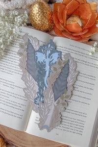This bookish When The Moon Hatched dragonscale dagger die-cut bookmark is inspired by the Moonfall series by Sarah A Parker. The image shows the bookmark on an open book, with the book placed on a fuzzy neutral colored blanket, a faux golden dragon egg, orange slices, and an orange lotus shaped tea light holder all surrounded by little white flowers