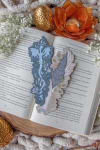 This bookish When The Moon Hatched dragonscale dagger die-cut bookmark is inspired by the Moonfall series by Sarah A Parker. The image shows the bookmark on an open book, with the book placed on a fuzzy neutral colored blanket, a faux golden dragon egg, orange slices, and an orange lotus shaped tea light holder all surrounded by little white flowers