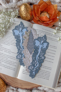 This bookish When The Moon Hatched dragonscale dagger die-cut bookmark is inspired by the Moonfall series by Sarah A Parker. The image shows the bookmark on an open book, with the book placed on a fuzzy neutral colored blanket, a faux golden dragon egg, orange slices, and an orange lotus shaped tea light holder all surrounded by little white flowers