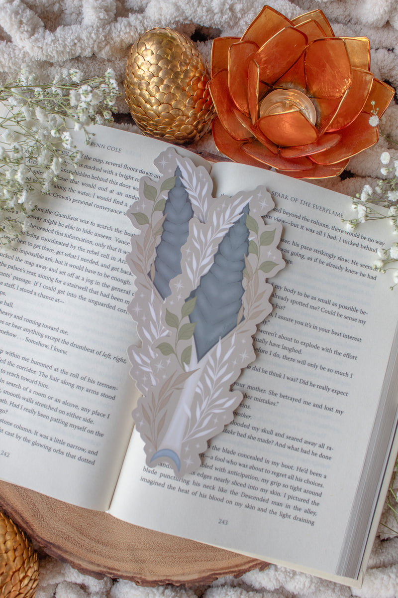 This bookish When The Moon Hatched dragonscale dagger die-cut bookmark is inspired by the Moonfall series by Sarah A Parker. The image shows the bookmark on an open book, with the book placed on a fuzzy neutral colored blanket, a faux golden dragon egg, orange slices, and an orange lotus shaped tea light holder all surrounded by little white flowers
