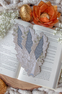 This bookish When The Moon Hatched dragonscale dagger die-cut bookmark is inspired by the Moonfall series by Sarah A Parker. The image shows the bookmark on an open book, with the book placed on a fuzzy neutral colored blanket, a faux golden dragon egg, orange slices, and an orange lotus shaped tea light holder all surrounded by little white flowers
