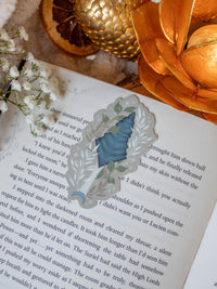 This bookish When The Moon Hatched dragonscale dagger sticker is inspired by the Moonfall series by Sarah A Parker. The image shows the sticker on an open book, with the book placed on a fuzzy neutral colored blanket, a faux golden dragon egg, orange slices, and an orange lotus shaped tea light holder all surrounded by little white flowers